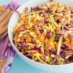 Horizontal image: white bowl with red and green cabbage coleslaw inside. Left of bowl: a purple kitchen towel with two wooden utensils on top.