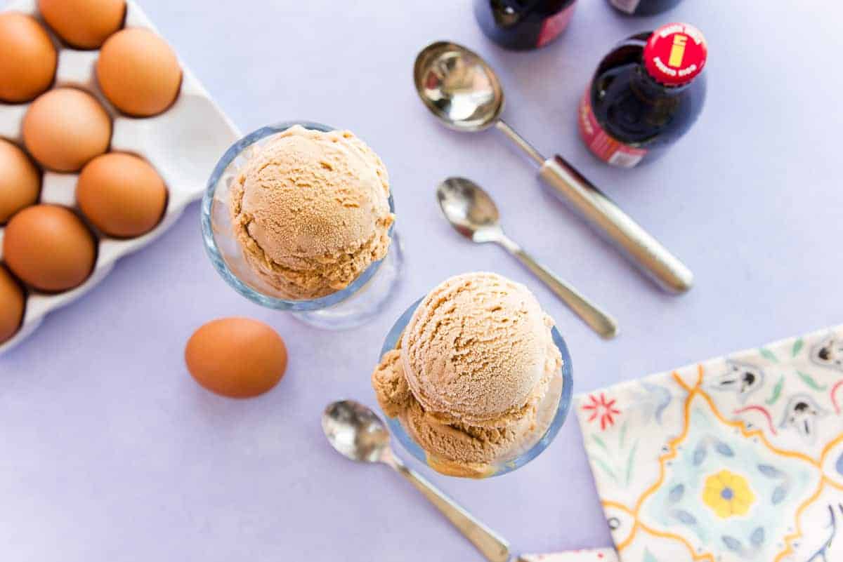 Overhead horizontal image of two tall glasses filled with scoops of Ponche Frozen Custard surrounded by brown eggs and bottles of Malta.