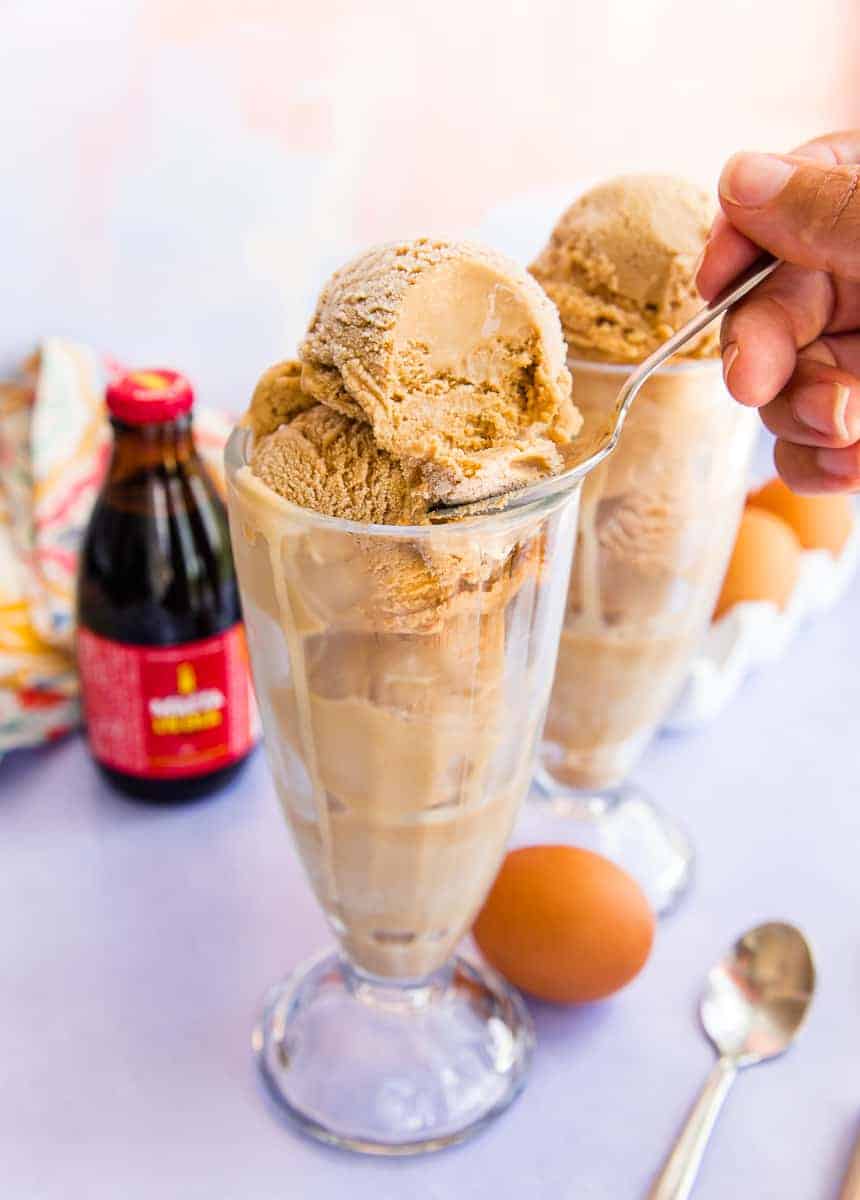 A hand scoops a spoonful of Ponche Frozen Custard from a top of scoop of custard in a clear milkshake glass. A brown egg is at the base of the glass and a brown bottle of Malta is in the left side of the image.