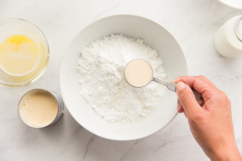 Evaporated milk is added to a white bowl of powdered sugar.