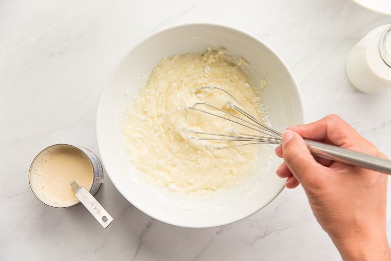A hand uses a silver whisk to stir the milks into powdered sugar in a white ceramic bowl.