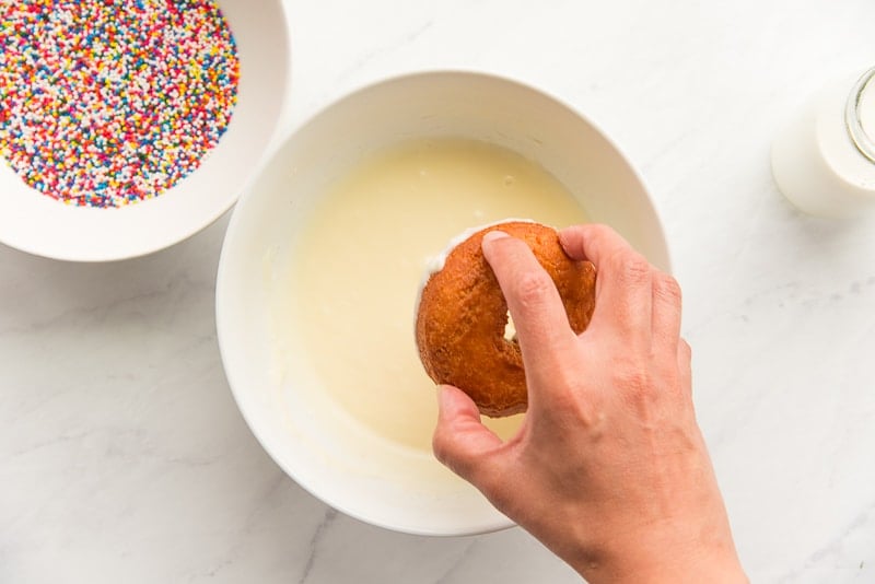 A hand pulls a cake donut from the milk glaze in a white ceramic bowl. A white bowl of rainbow sprinkles is in top left corner.
