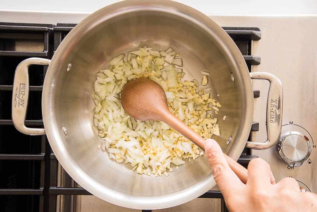 Hand uses a spoon to stir sauteing onions and garlic in a silver pot on a stove top.