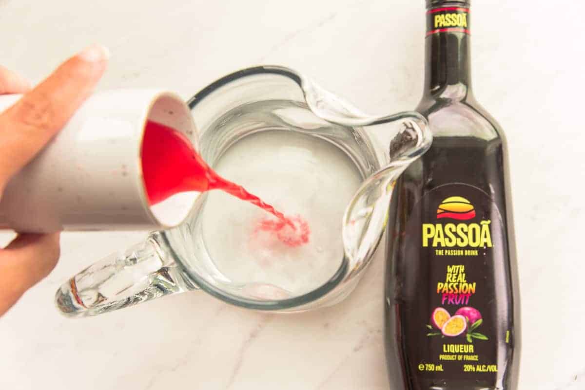 Passoa is poured from a small white pitcher into a clear glass pitcher of simple syrup.