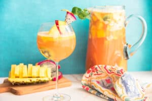 Preview image of a wine goblet of Tropical Sangria next to a wooden cutting board with pineapple and dragonfruit and a glass pitcher of sangria.