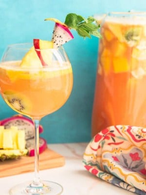 Preview image of a wine goblet of Tropical Sangria next to a wooden cutting board with pineapple and dragonfruit and a glass pitcher of sangria.