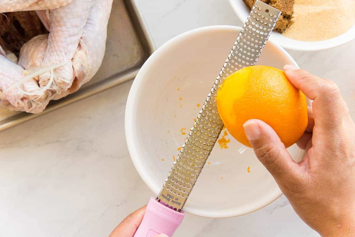 A hand uses a microplane to zest the peel of an orange into a white mixing bowl