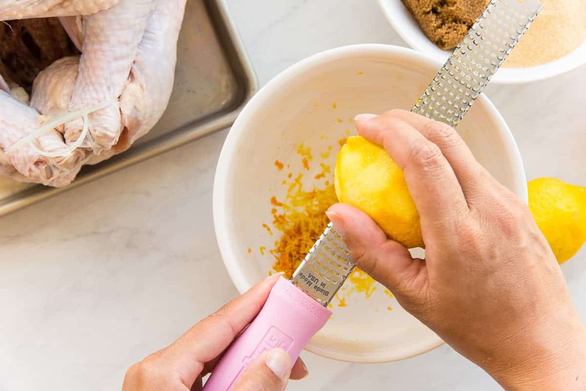 A hand uses a microplane to zest the peel of a lemon into a white bowl
