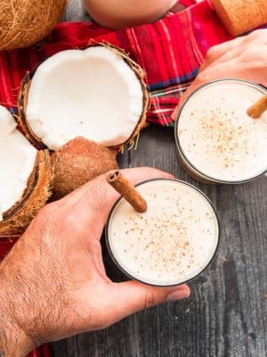 Coquito Recipes preview, two hands toasting with two glasses of coquito garnished with cinnamon sticks.