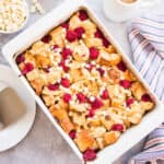 Overhead portrait image of a white dish of Raspberry White Chocolate Bread Pudding surrounded by bowls of raspberries, white chocolate chips and a pitcher of sauce