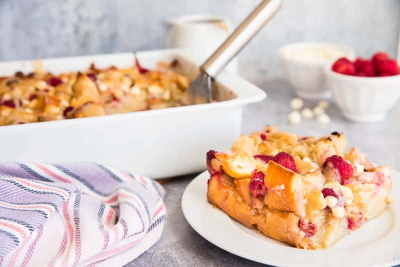 Horizontal, close up of a serving of Raspberry White Chocolate Bread Pudding on a small white plate. A white bowl of raspberries in right background