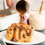 Coquito Toffee Sauce is poured from a white pitcher onto a serving of Coquito Bread Pudding on a white plate.