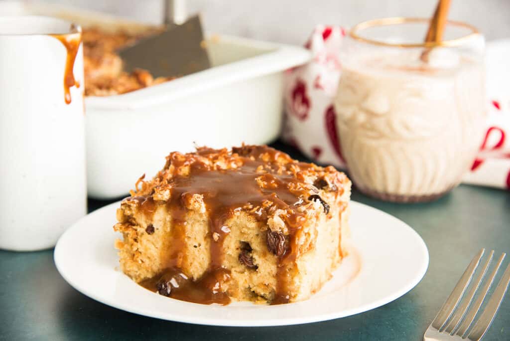Horizontal image: square serving of Coquito Bread Pudding on a white plate in front of a santa cocktail glass of coquito.