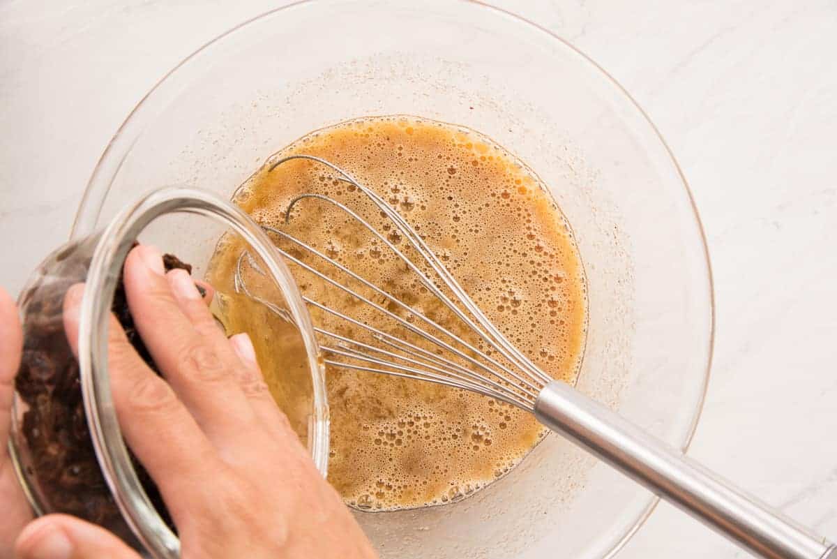 The soaking liquid from the raisins is poured into the egg and sugar mixture in a clear, glass mixing bowl.