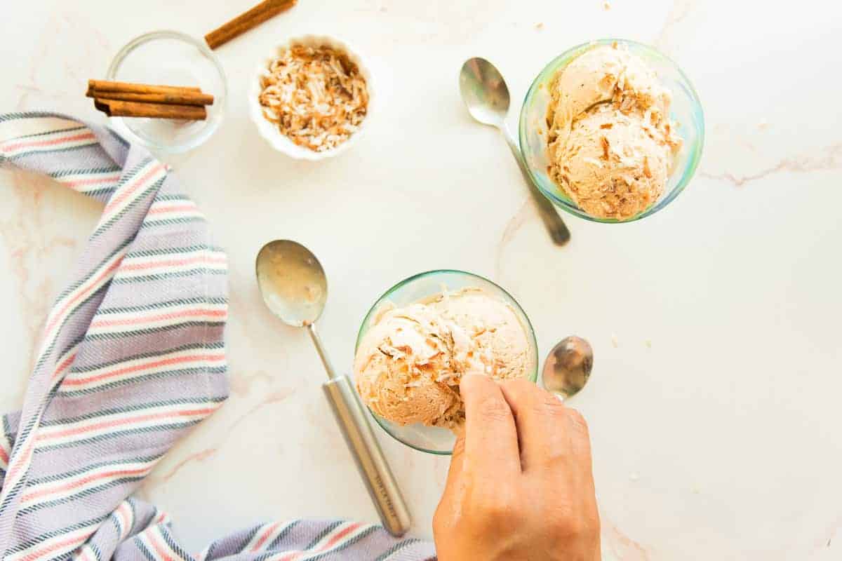 A hand sprinkles toasted coconut flakes on top of scoops of Coquito Frozen Custard in a green ice cream dish.