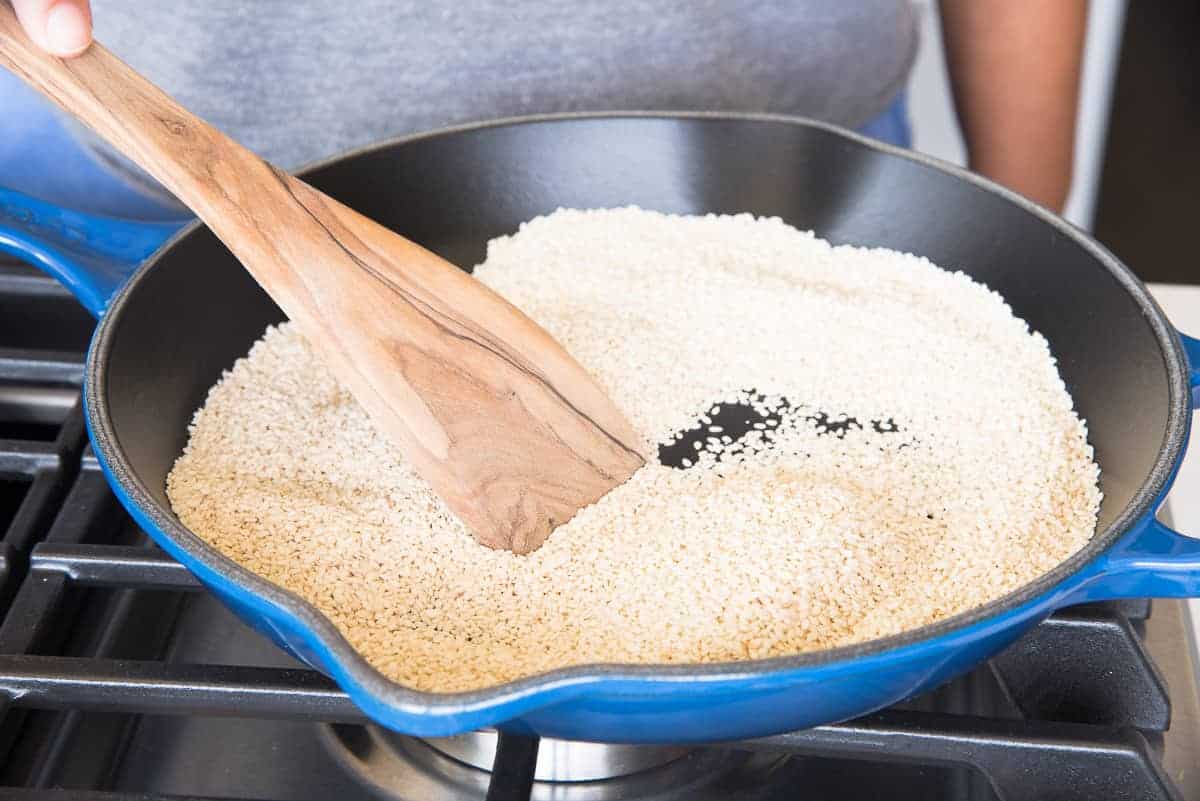 Raw, white sesame seeds are added to pan and stirred with a flat wooden spoon.