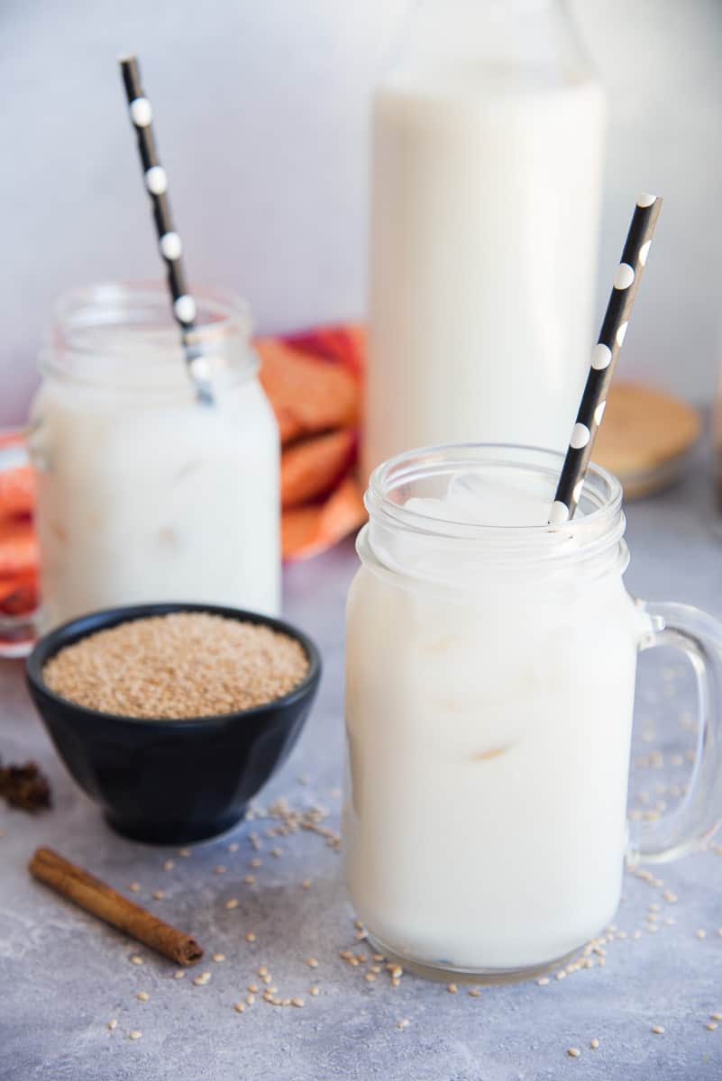 Portrait image of two clear mugs of Horchata de Ajonjolí with black and white polka dot straws next to a bowl of toasted sesame seeds.