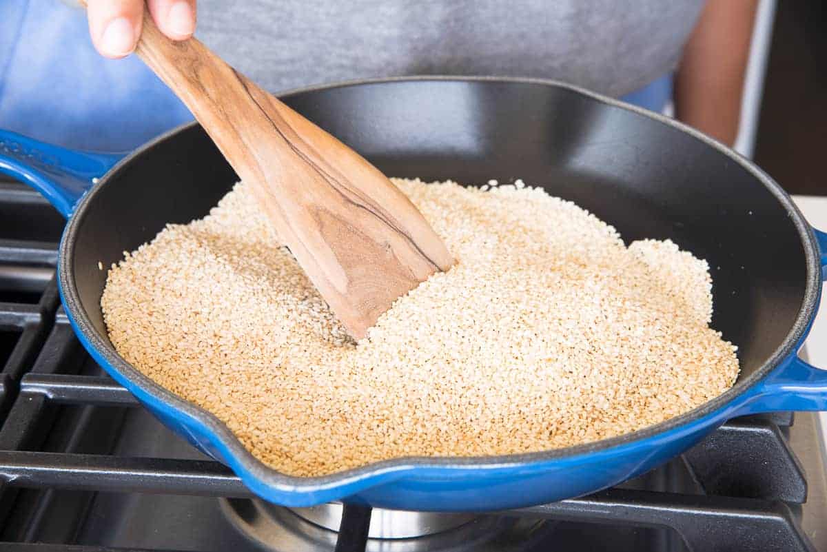 The raw sesame seeds are stirred with a wooden spoon in a blue and black pan.