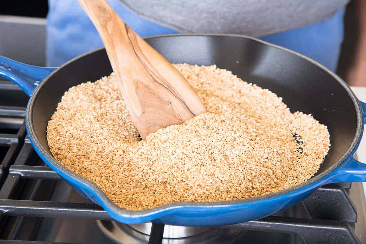 A wooden spoon stirs the sesame seeds as they are almost done toasting in a blue pan.