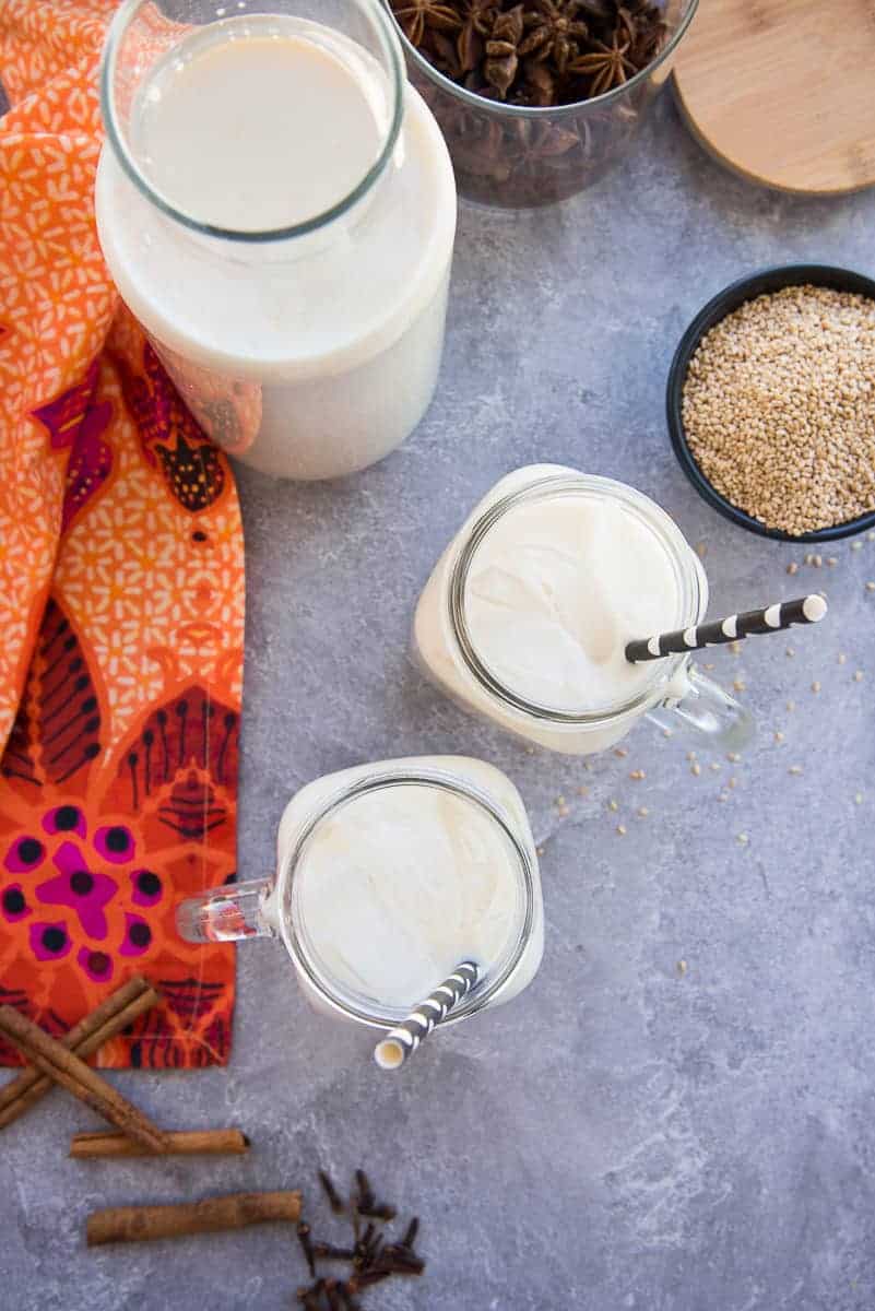 Portrait image of a carafe of Sesame Seed Milk (Horchata de Ajonjolí) next to two glass mugs of horchata and a bowl of toasted sesame seeds.