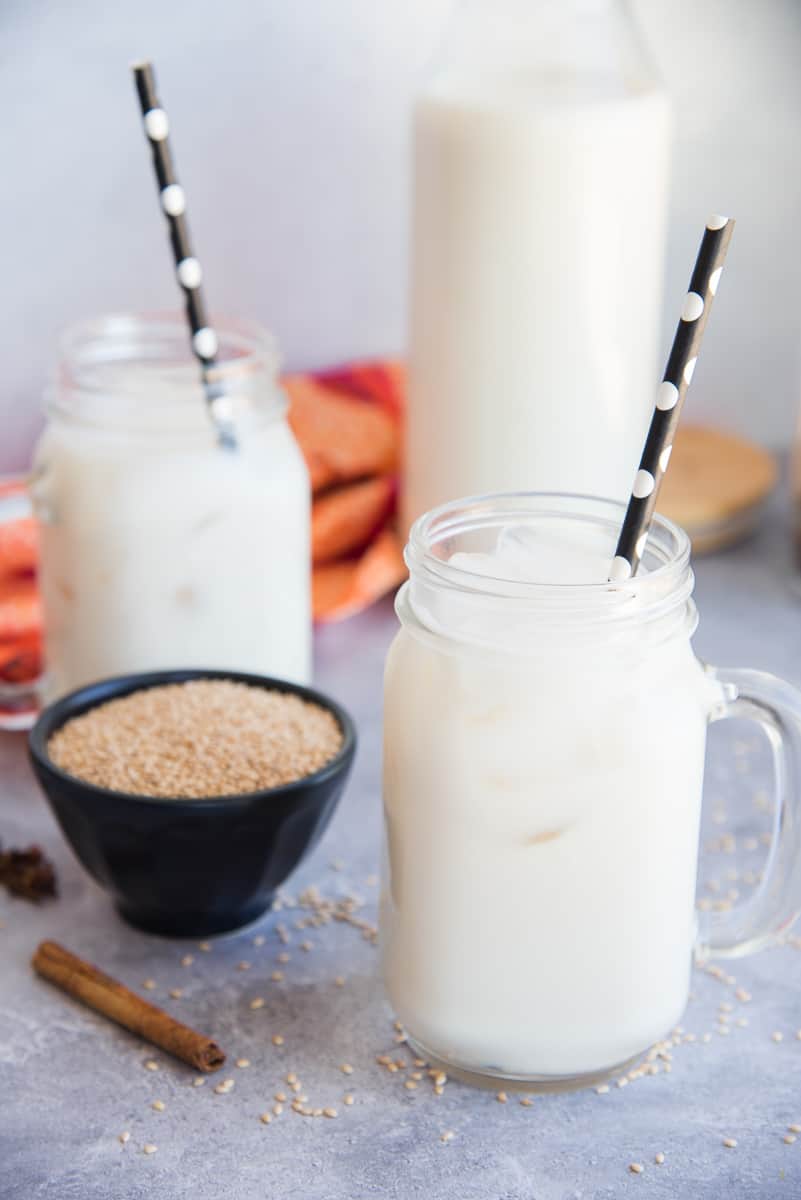 Lead image of two glass mugs of sesame seed milk horchata de ajonjolí with black and white polka dot straws in front of a carafe of sesame seed milk