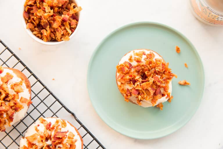 A bagel topped with cream cheese and crispy bacon pieces sits on a green plate. Nearby, more bagels with the same toppings rest on a cooling rack, and a small container holds additional bacon. A light-colored surface serves as the backdrop.