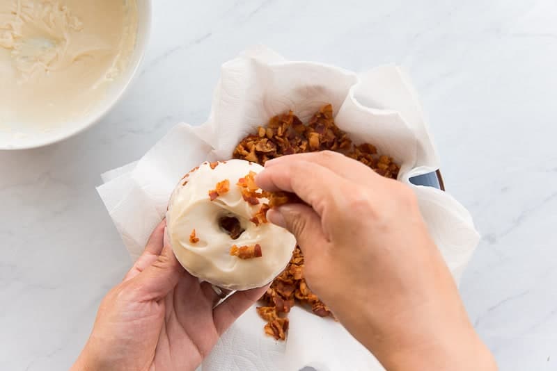 Hand holds the maple-glazed donut while the other hand sprinkles cooked bacon bits on top of it.
