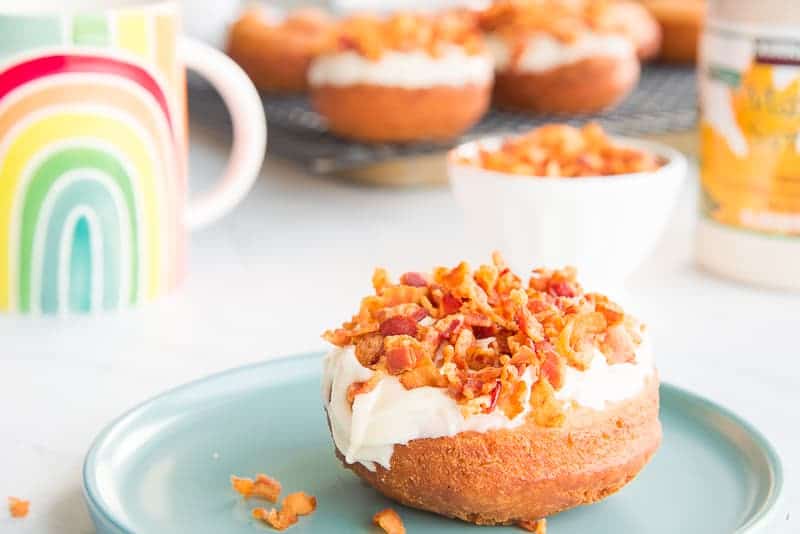 A maple bacon donut on a teal plate in front of a rack with more maple bacon donuts on it.