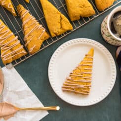Preview image of a silver cooling rack with Pumpkin scones on them next to a glass bowl of Maple Cinnamon Cream Cheese Glaze and a white plate with one scone on it.