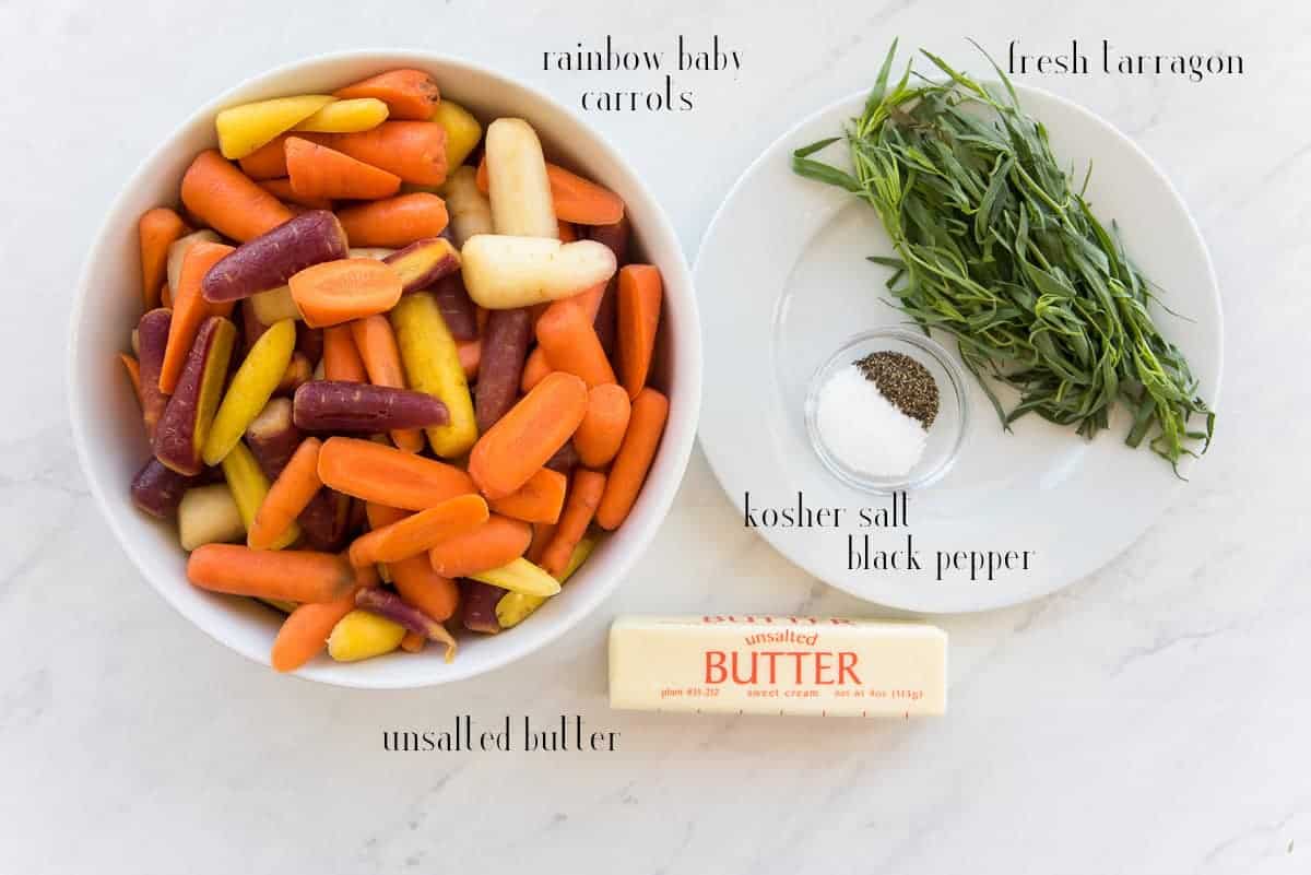 The ingredients to make Rainbow Carrots in Tarragon Brown Butter Sauce on a white surface: rainbow carrots, fresh tarragon, kosher salt, black pepper, and unsalted butter
