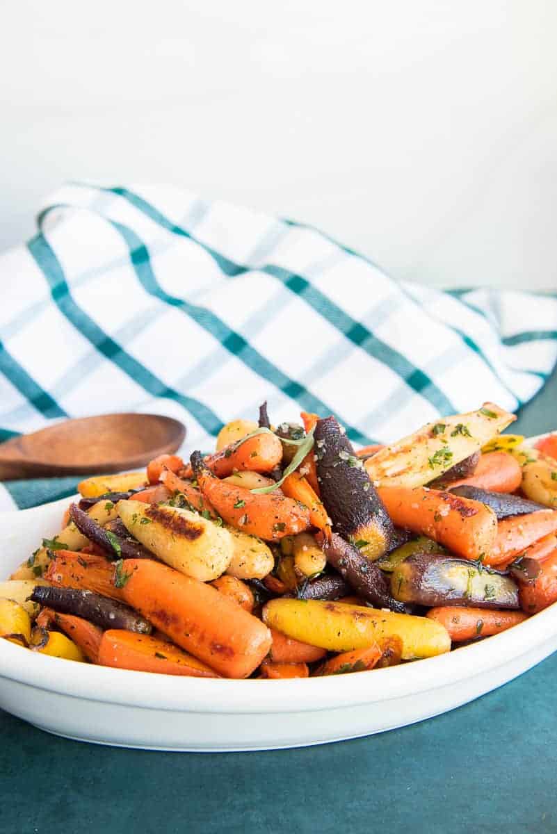 Portrait image of a white dish of Rainbow Carrots in Tarragon Brown Butter Sauce garnished with tarragon leaves.