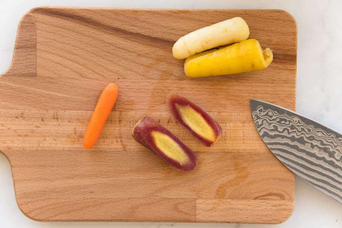 Four different colored rainbow baby carrots are on a light-colored wooden board. A purple carrot is cut in half diagonally. An orange carrot, which is too thin to cut, is left whole.