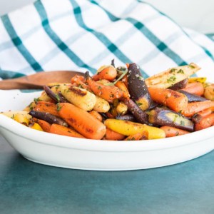 Preview image of a white, oblong bowl of Rainbow Carrots in Tarragon Brown Butter Sauce on a dark green surface.