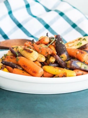 Preview image of a white, oblong bowl of Rainbow Carrots in Tarragon Brown Butter Sauce on a dark green surface.