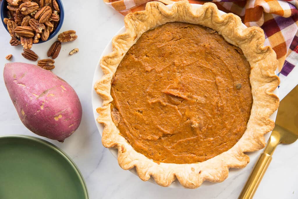 A freshly baked sweet potato pie with a crimped crust sits on a white surface. Nearby are a whole sweet potato, a bowl of pecans, a green plate, and a golden serving spatula. A checkered cloth with orange and purple hues is partially visible in the background.