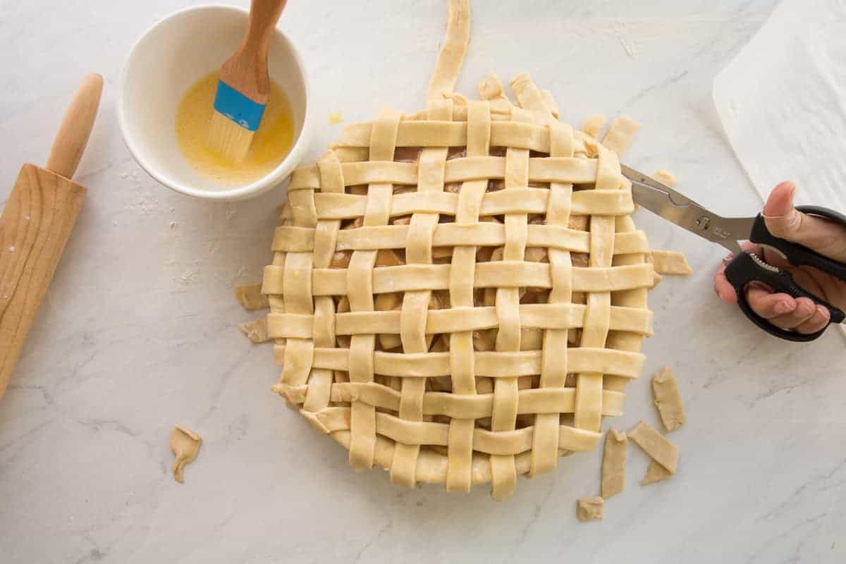 A hand uses kitchen shears to trim the excess pie dough from the lattice top