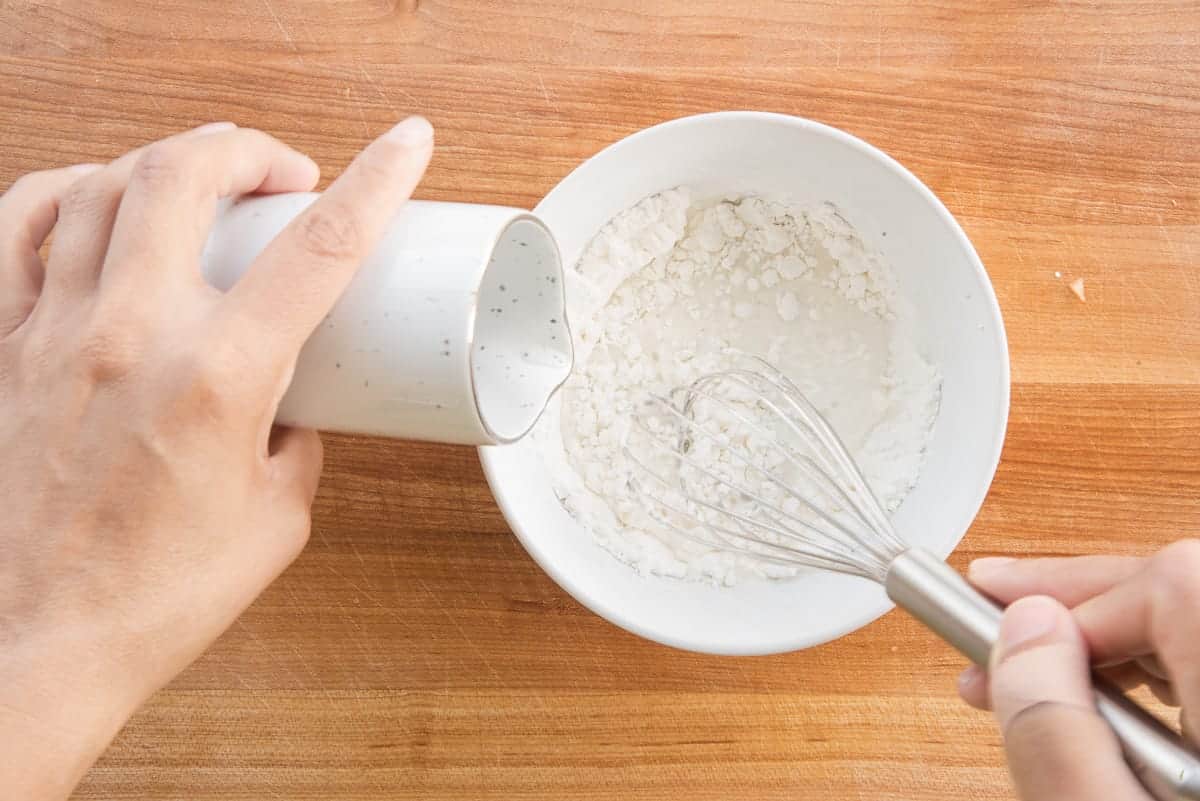 A hand pour water from a white pitcher into a bowl of cornstarch and whisks the two together.
