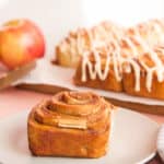 Social media image of a single, unglazed Apple Cinnamon Roll on a light grey plate in front of a batch of rolls drizzled in Apple Butter Glaze