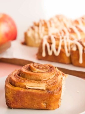Preview image of a single Apple Cinnamon Roll on a light grey plate in front of the remaining rolls drizzled in Apple Butter Glaze.
