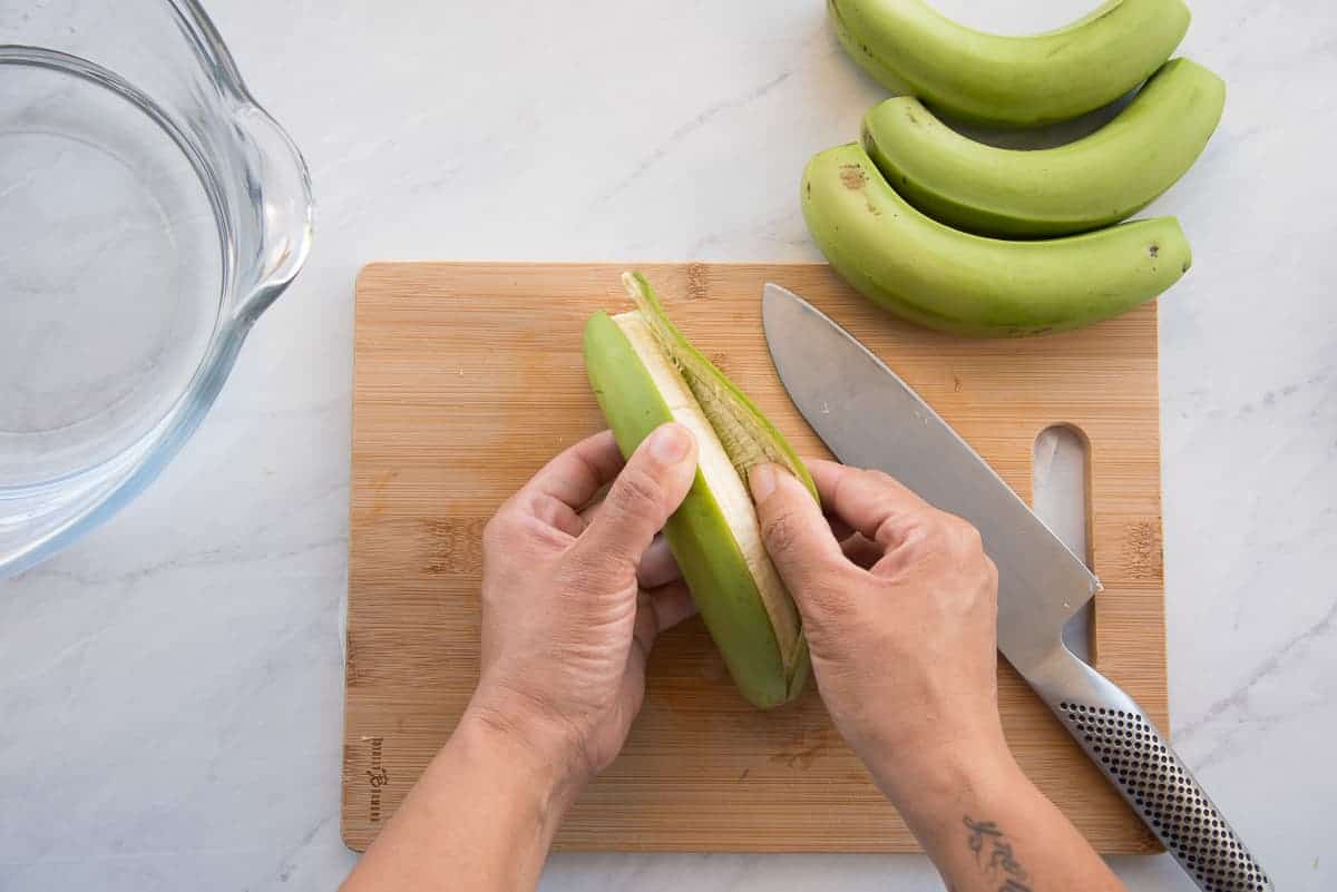 Two hands peel the skin from a green cooking banana.