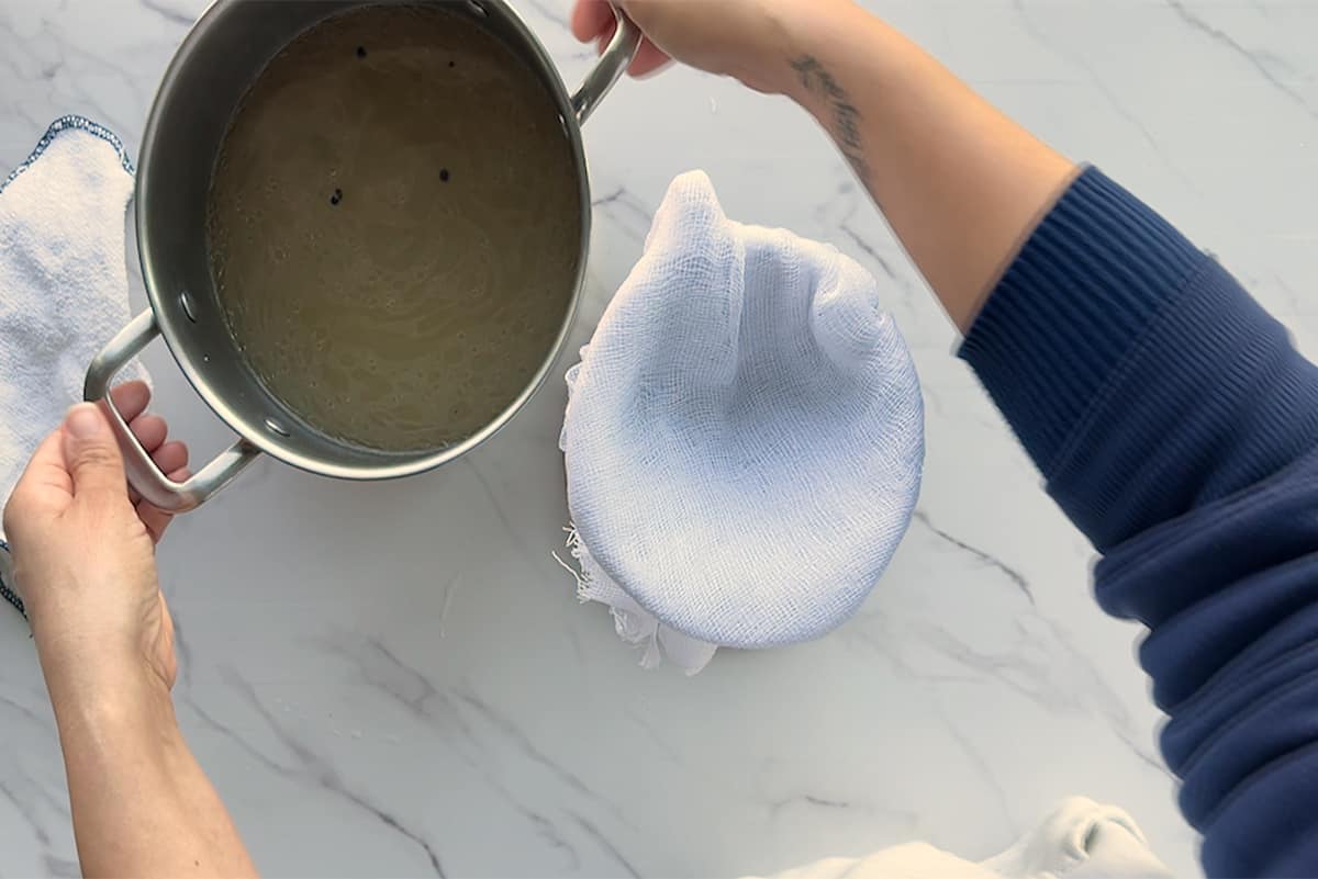 Hands lift the stock pot to pour the ham broth into a cheesecloth strainer.