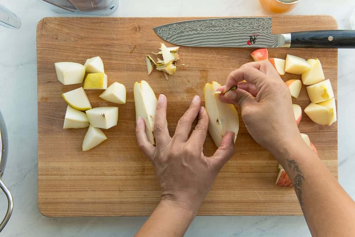 Hands remove the cores and stems from a pear. The fruits are chopped on a wooden cutting board.