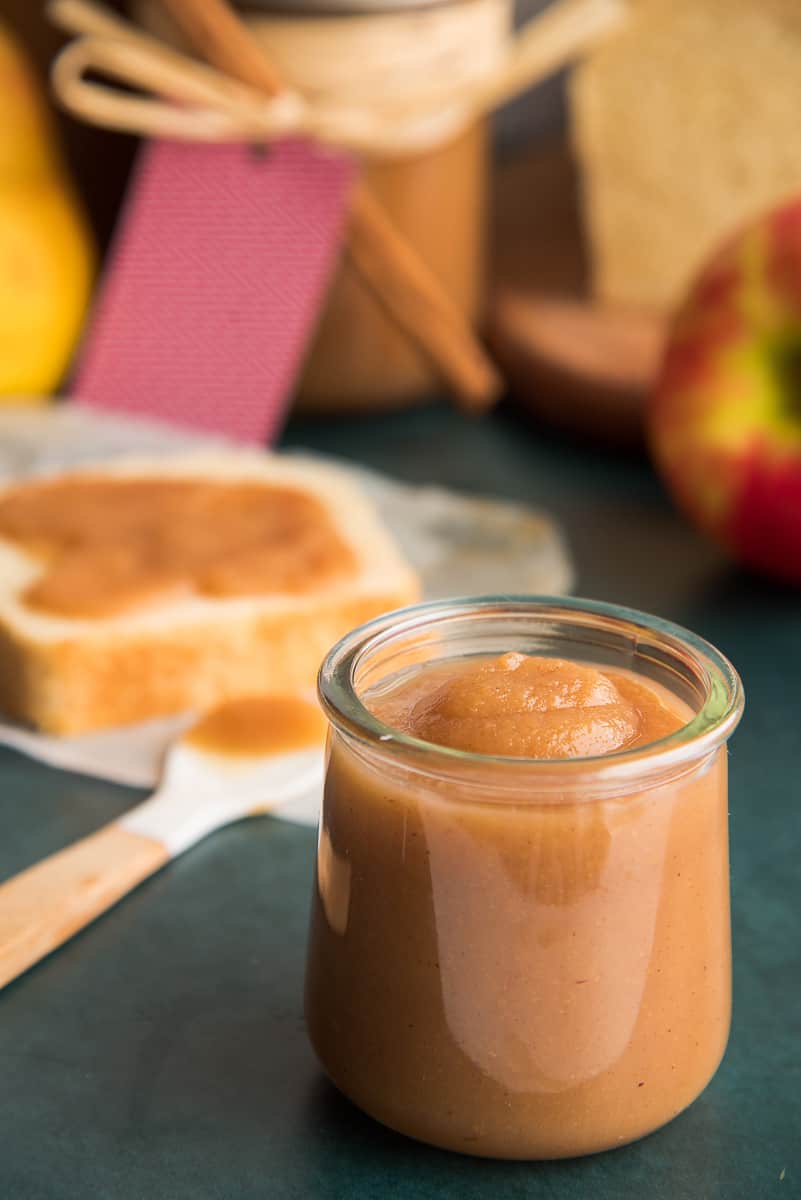 Portrait image of a glass jar of Slow Cooker Apple Pear Butter in front of a slice of bread spread with the butter