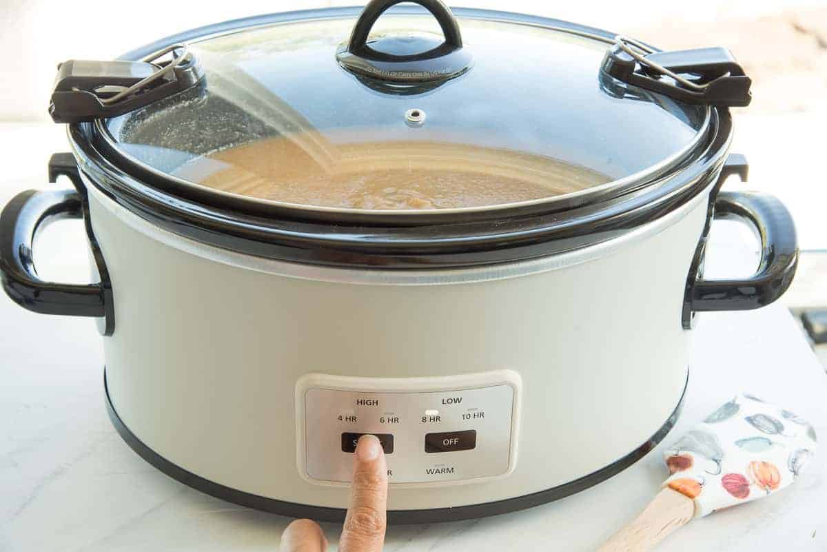 A finger presses the temperature button on a white slow cooker to begin cooking the fruit mixture.