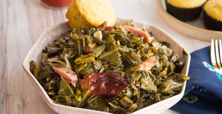Horizontal image of a pink bowl of slow cooker collard greens in front of a bottle of red hot sauce.