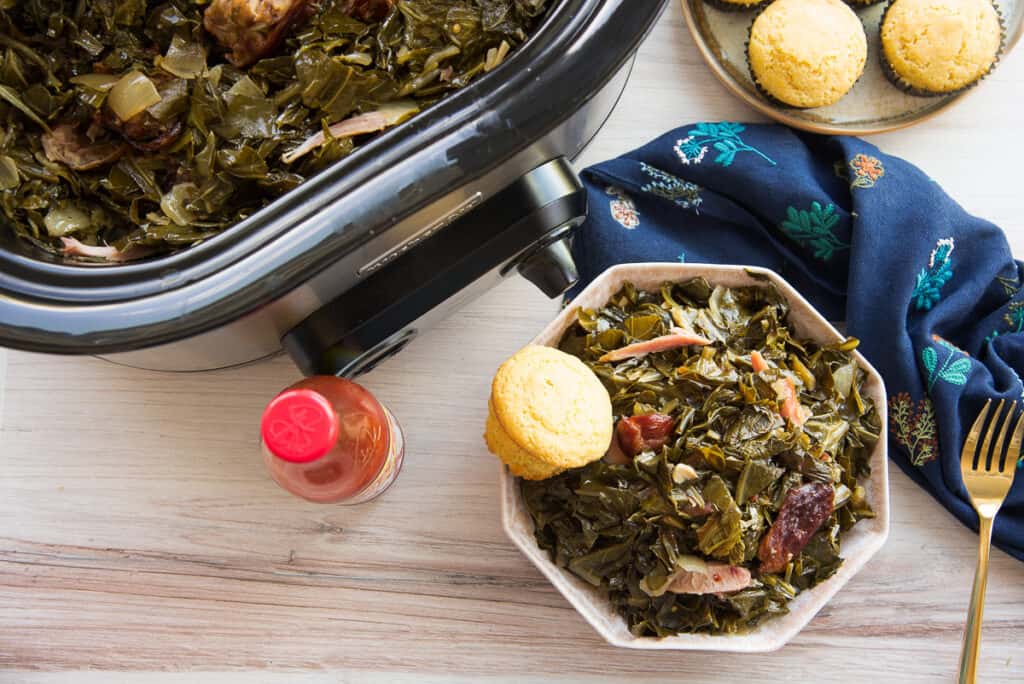 Overhead horizontal image of a pink hexagonal bowl of country style slow cooker collard greens with a cornbread muffins next to a red capped hot sauce bottle.