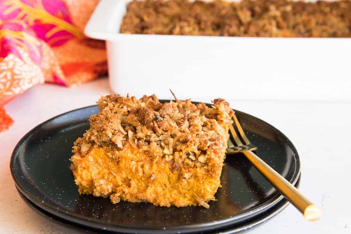 Horizontal image shows a cut piece of sweet potato casserole with coconut pecan topping on two stacked black plates with white specks. A gold fork is on the right of the serving of casserole on the plate.