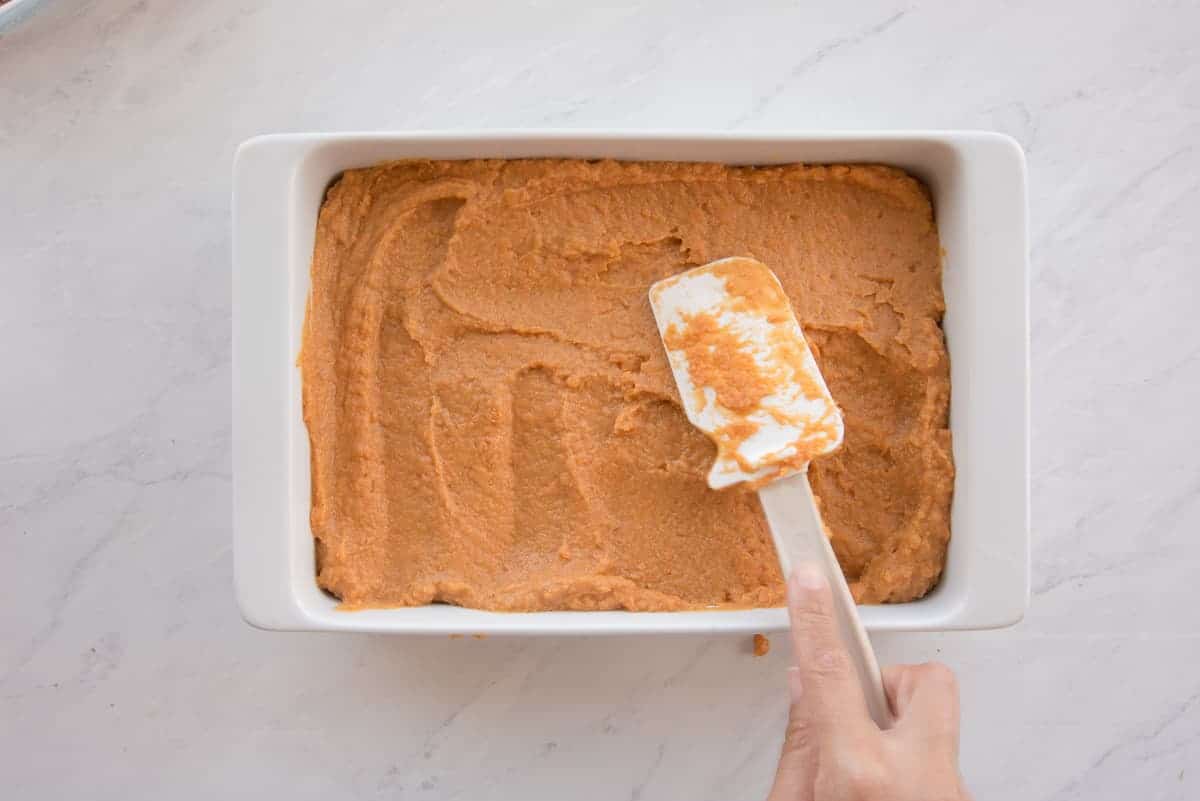 A hand uses a white rubber spatula to smooth the sweet potato mixture into a white ceramic baking dish.