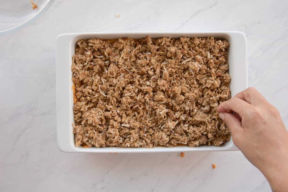 A hand sprinkles the coconut pecan topping on top of the sweet potato mixture in a white ceramic baking dish.