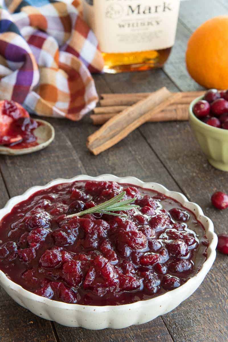 Portrait image of a cream bowl filled with Boozy Spiced Cranberry Sauce garnished with a sprig of rosemary. The rest of the recipe ingredients are in the background.